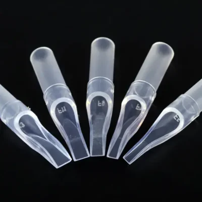 Five clear disposable plastic tattoo tips fanned out — flat magnum openings with size markings and transparent cylindrical grip sleeves