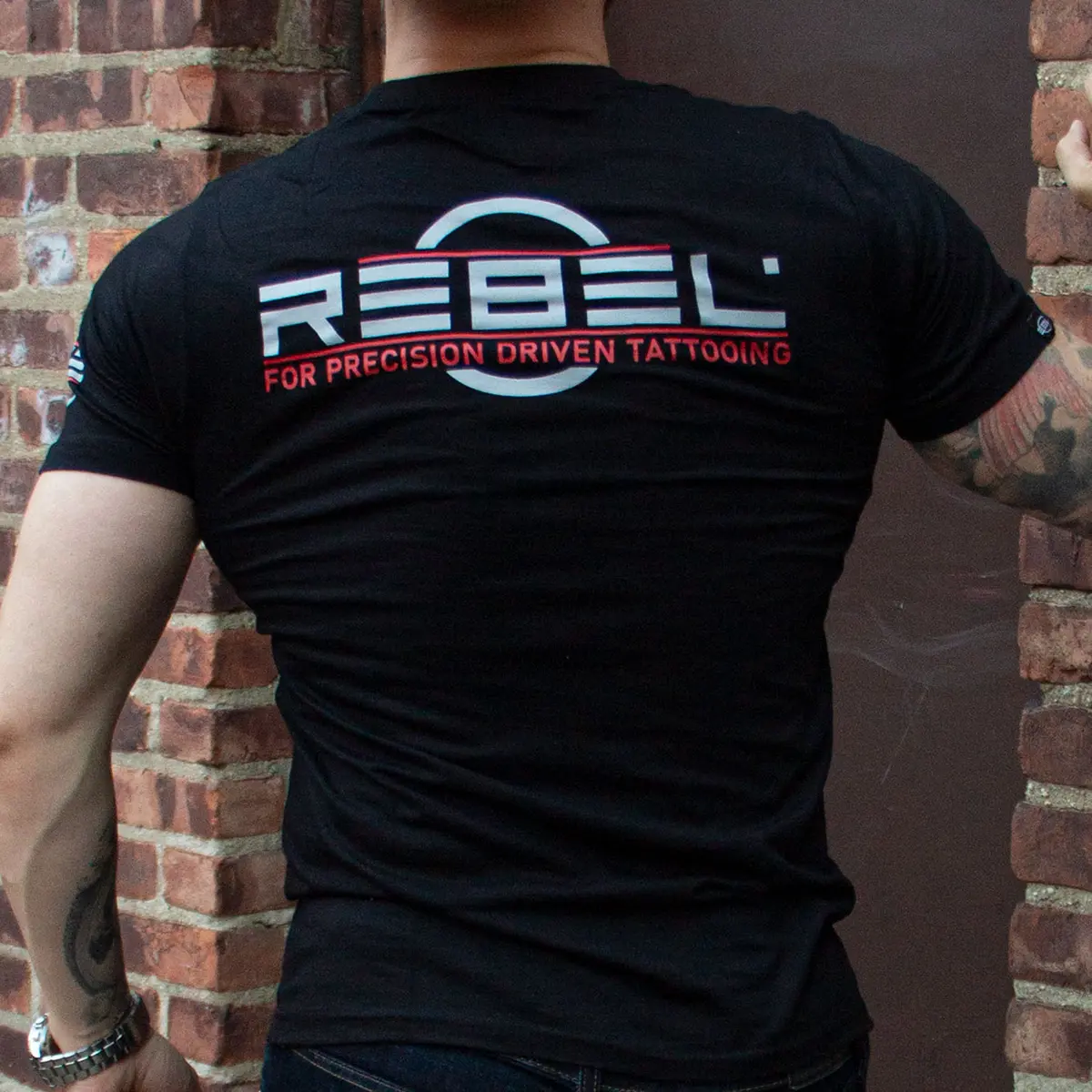 Male model wearing REBEL Precision Tee back view, REBEL FOR PRECISION DRIVEN TATTOOING slogan, brick wall