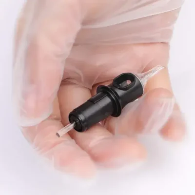 Tattoo cartridge held in gloved hand showing needle tip and grip interface, cartridge housing and plunger stem