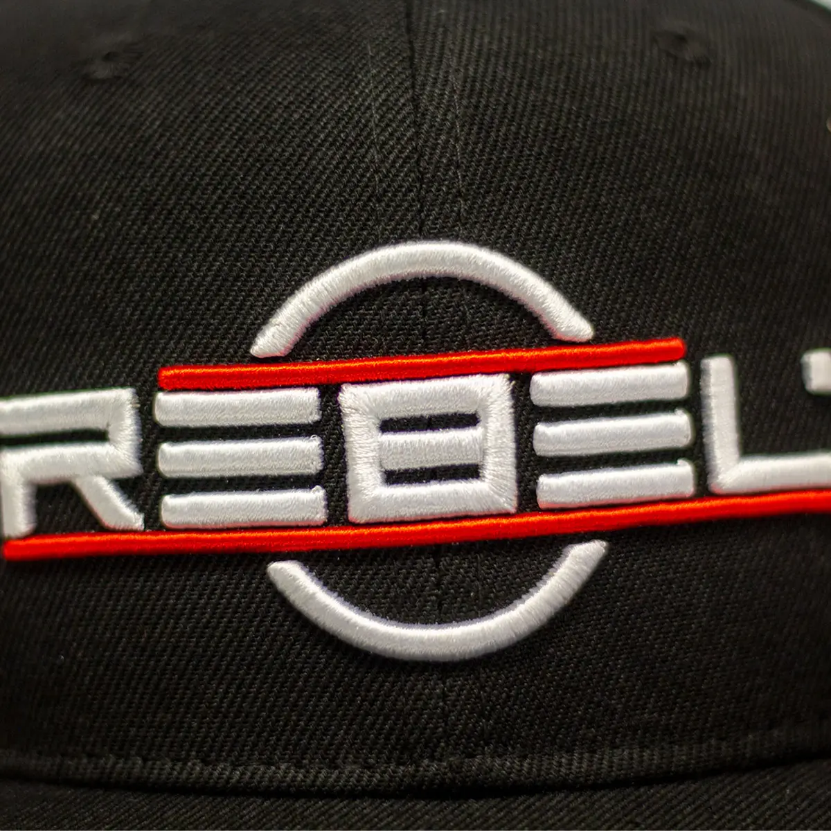 Close-up of the 3D white and red REBEL embroidery on the front panel of the black snapback hat showing raised thread detail and circle logo
