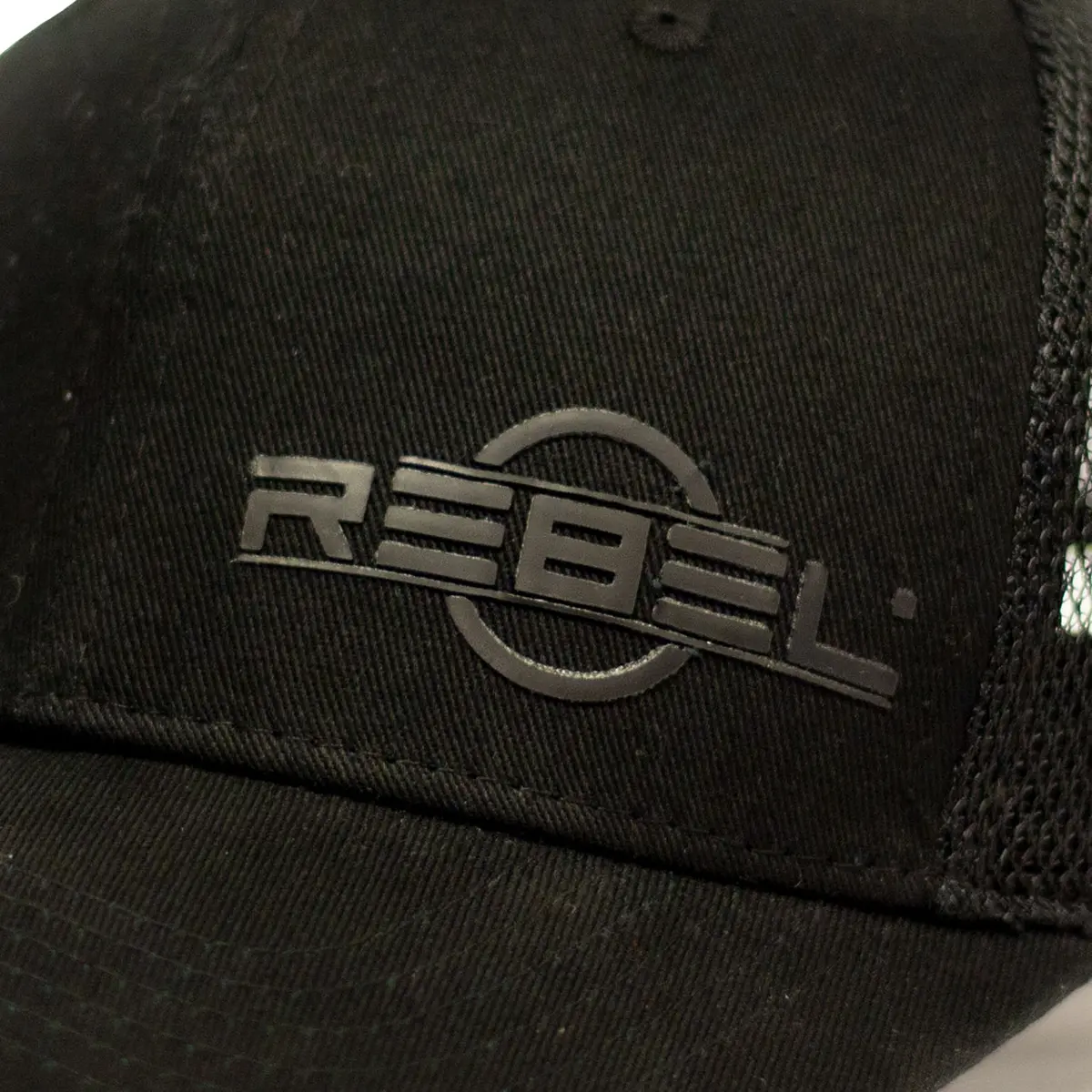 Close-up of raised black rubber REBEL logo on structured cotton front panel of the REBEL Phantom Trucker Hat