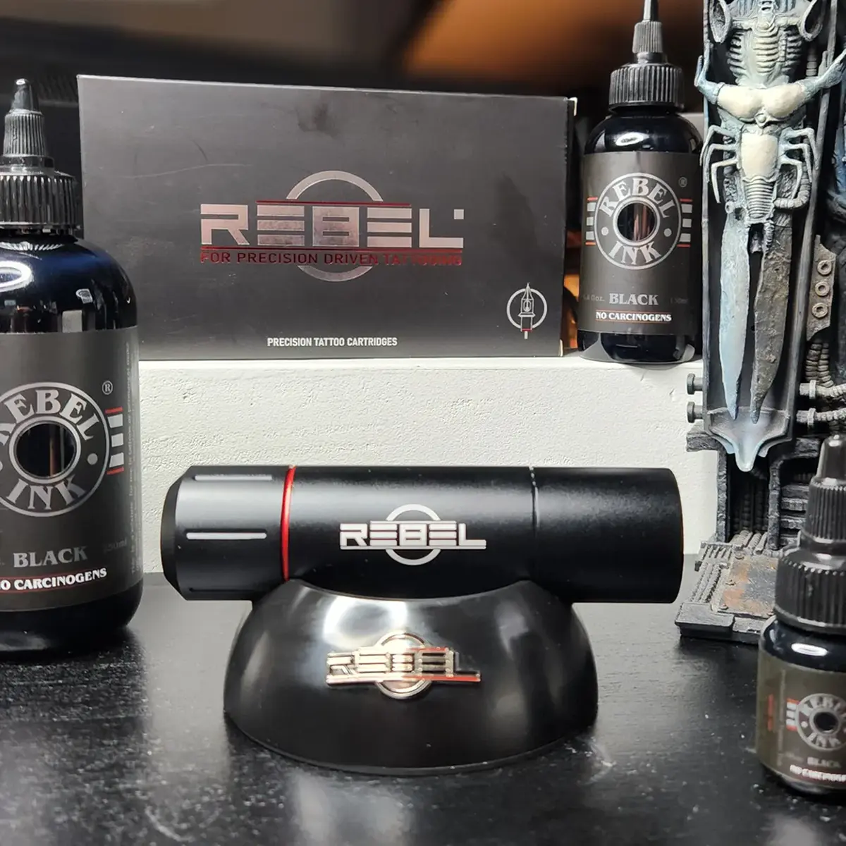 Tattoo machine holder on studio desk with REBEL Ink bottles and Precision Cartridges box, with prop tattoo machine
